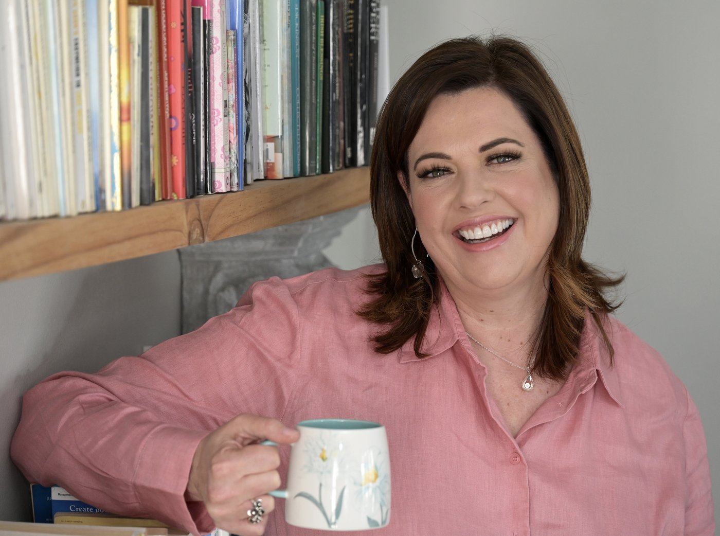 Zeldi laughing warmly, holding a mug, standing in front of a bookshelf.